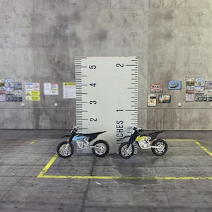 Könnte beinhalten: Zwei Miniatur-Motocross-Motorräder, eines hellblau-schwarz, das andere gelb-schwarz. Ein Lineal zeigt, dass die Motorräder etwa 2,5 cm lang sind. Sie stehen auf einer grauen Oberfläche.