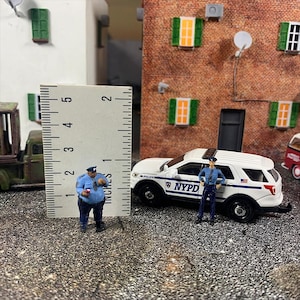 Puede incluir: Escena en miniatura con una figura de policía y un SUV blanco de la policía de Nueva York. El oficial, sosteniendo una taza, está de pie junto a una regla, midiendo aproximadamente 2,5 cm. Un edificio de ladrillo con contraventanas verdes es el telón de fondo.