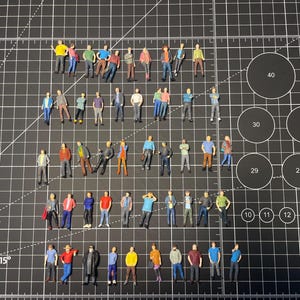 May include: Miniature figures of people in various poses and outfits, including jackets, shirts, and pants. The figures are in different colors and are arranged on a black grid surface. The figures are small, likely for a model or diorama.