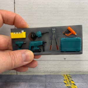 Op de afbeelding: Een set miniatuur gereedschap, waaronder een boormachine, een hamerboor, een cirkelzaag, een tang, een nietpistool, een gereedschapskist en een gele batterijpack. De gereedschappen zijn gerangschikt op een grijze achtergrond.