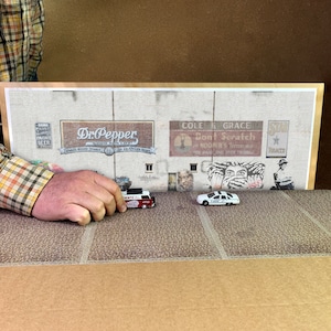 Könnte beinhalten: Eine weiße Backsteinwand mit einem Wandbild mit Vintage-Werbeanzeigen für Dr Pepper, Cole & Grace und Star Tobacco. Das Wandbild ist im Retro-Stil mit kräftigen Farben und Schriftzügen gemalt. Vor der Wand stehen zwei Spielzeugautos, ein roter und weißer Van und ein weißes Polizeiauto.