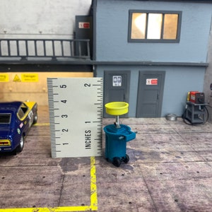 Könnte beinhalten: Ein blauer und gelber Ölwechselständer mit einem gelben Tablett oben. Der Ständer steht auf einer grauen Oberfläche mit einem Lineal im Hintergrund. Das Lineal ist in Zoll eingeteilt.