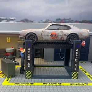 Può includere: Una scena di garage in miniatura con un'auto usurata su un ponte sollevatore. L'auto ha il logo "GULF". Una piccola figura di un meccanico sta lavorando sull'auto. Il ponte sollevatore ha la scritta "GEARMIGHT" sul lato.