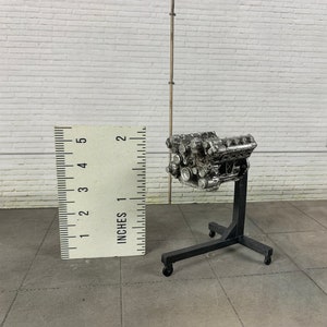 Può includere: Un modello argentato di un motore di auto su un supporto in metallo nero con ruote. Il motore è alto circa 5 cm e il supporto circa 10 cm.