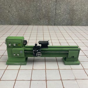 Puede incluir: Un torno miniatura de metal verde con una cabezal y una contrapunta de metal negro. El torno está sobre una superficie de azulejos blancos.