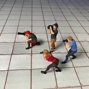 May include: Four miniature figures of photographers, each holding a camera and posed in different positions. The figures are standing on a white tiled surface.