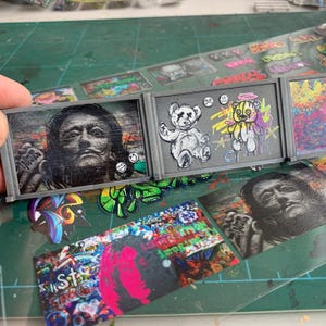 May include: Miniature art pieces in grey frames, including a portrait of Salvador Dalí and graffiti-style teddy bears. Additional sticker-like art elements are visible, with a focus on vibrant colours and urban art themes.
