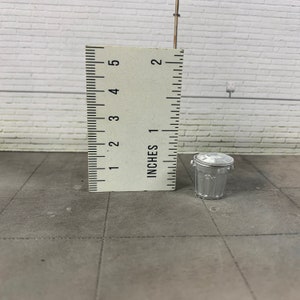 Puede incluir: Un cubo de basura de metal plateado con tapa, que mide aproximadamente 2,5 cm de alto. El cubo de basura está sobre una superficie de baldosas grises.