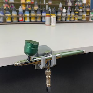 Peut inclure: Un aérographe vert et argenté est maintenu dans un support gris. L'aérographe a une coupelle verte et une buse argentée. Le support est fixé à une table. Une collection de flacons de peinture est en arrière-plan.