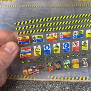 Puede incluir: Una hoja de calcomanías en miniatura con varios letreros de advertencia, incluyendo "Peligro", "Precaución" y "GLP". También se incluyen logotipos de estilo vintage para marcas como Shell, Castrol y Champion. Las calcomanías están sobre un fondo gris con rayas de peligro amarillas y negras.