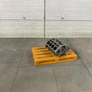 Könnte beinhalten: Ein silbernes Modell eines großen Motorblocks, der auf einer kleinen Holzpalette steht. Die Palette besteht aus hellbraunem Holz und hat ein paar Latten.