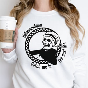 May include: White sweatshirt featuring a black and white graphic of a skeleton wearing a hat, with the words "Halloweentown" and "Catch me in the next life" in a circular design. A white coffee cup with a brown sleeve is held in the left hand.