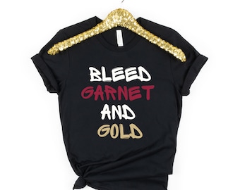 May include: Black t-shirt with the text "BLEED GARNET AND GOLD" in white, red, and gold lettering.