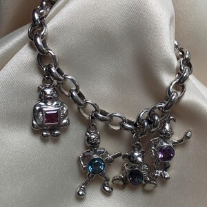 Peut inclure: Bracelet à breloques en argent avec quatre breloques fantaisistes : un ours avec une gemme carrée rose, une figurine avec une gemme bleue, un ours assis avec une gemme foncée et un lapin avec une gemme violette. Le bracelet a un design de chaîne épaisse.
