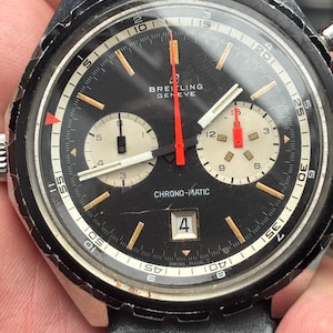 Könnte beinhalten: Vintage Breitling Chrono-Matic Uhr mit schwarzem Zifferblatt, zwei Hilfszifferblättern und Datumsfenster bei 4 Uhr. Die Uhr hat ein Edelstahlgehäuse, ein schwarzes Lederarmband und rote und weiße Akzente. Das Zifferblatt hat einen Durchmesser von ca. 3,8 cm.