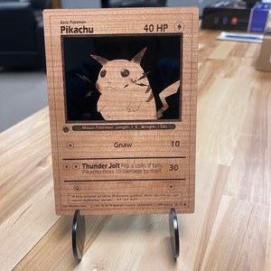 May include: A wooden Pokemon trading card featuring Pikachu, a mouse Pokemon. The card has a black background with a picture of Pikachu in the center. The card has the text "Basic Pokemon Pikachu 40 HP" at the top and "Gnaw 10 Thunder Jolt Flip a coin, If tails, Pikachu does 10 damage to itself. 30" at the bottom.
