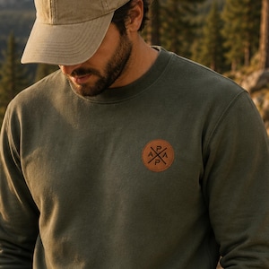 Peut inclure: Un sweat-shirt vert olive avec un écusson rond marron portant un logo. Le sweat-shirt est associé à une casquette de baseball kaki et à un jean foncé. L'image est prise en extérieur avec un arrière-plan montagneux.