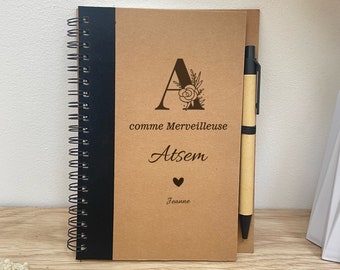 May include: A brown notebook with a black spiral binding and a black pen. The notebook has a floral design with the letter "A" and the text "comme Merveilleuse Atsem Jeanne".