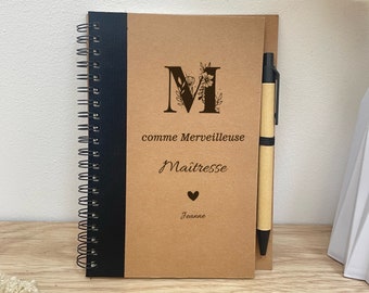 May include: A brown notebook with a black spiral binding and a black pen. The cover has a floral design with the letter "M" and the text "comme Merveilleuse Maitresse Jeanne".