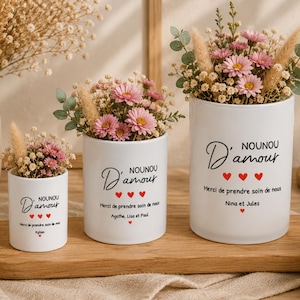 Peut inclure: Trois vases en céramique blanche avec des compositions florales. Les vases portent l'inscription "NOUNOU D'amour" et un message avec des cœurs rouges. Les fleurs sont roses et blanches, avec de la verdure et des herbes sèches.