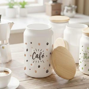 Peut inclure: Plusieurs pots en céramique blanche avec des couvercles en bois. L'un d'eux porte l'inscription "Café" avec des grains de café. Un autre est marqué "Thé". Une cafetière et une tasse de café sont également visibles.