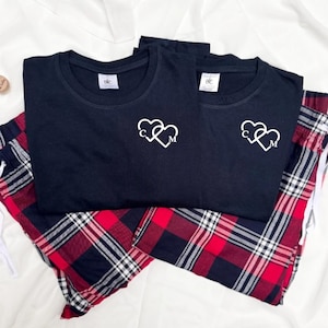 Puede incluir: Dos camisetas negras con iniciales blancas en forma de corazón, combinadas con pantalones de pijama a cuadros rojos y negros. Las camisetas tienen cuello redondo y un pequeño logo. Los pantalones de pijama tienen cintura con cordón.