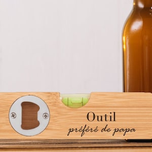 Könnte beinhalten: Ein Holz-Wasserwaage mit einem Flaschenöffner und einer grünen Libelle. Die Wasserwaage ist mit dem Text "Outil préféré de papa" graviert.