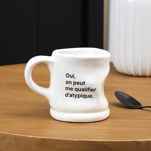 May include: White ceramic mug with an irregular shape and a black inscription that reads "Oui, on peut me qualifier d'atypique."