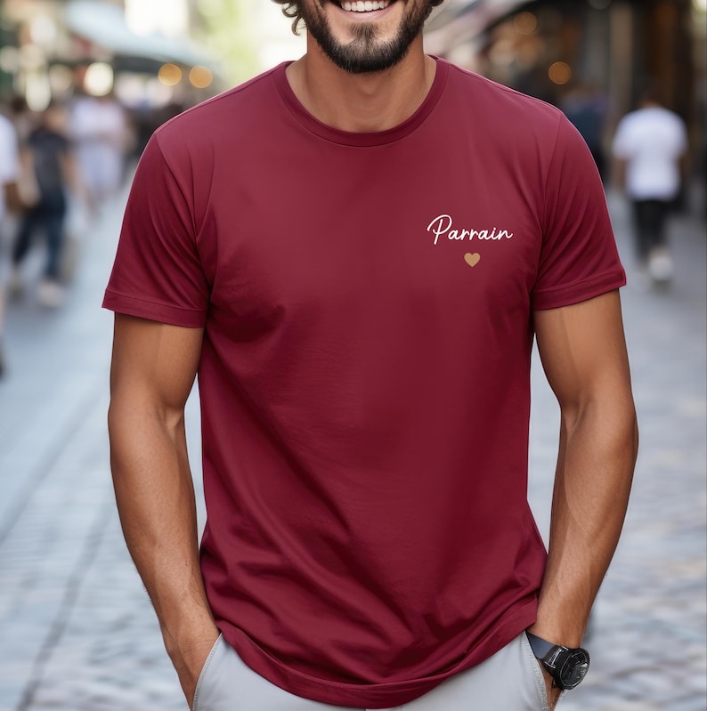 Puede incluir: Una camiseta granate con la palabra "Parrain" en escritura cursiva y un peque&ntilde;o coraz&oacute;n dorado. La camiseta es de manga corta y parece estar hecha de un material suave.