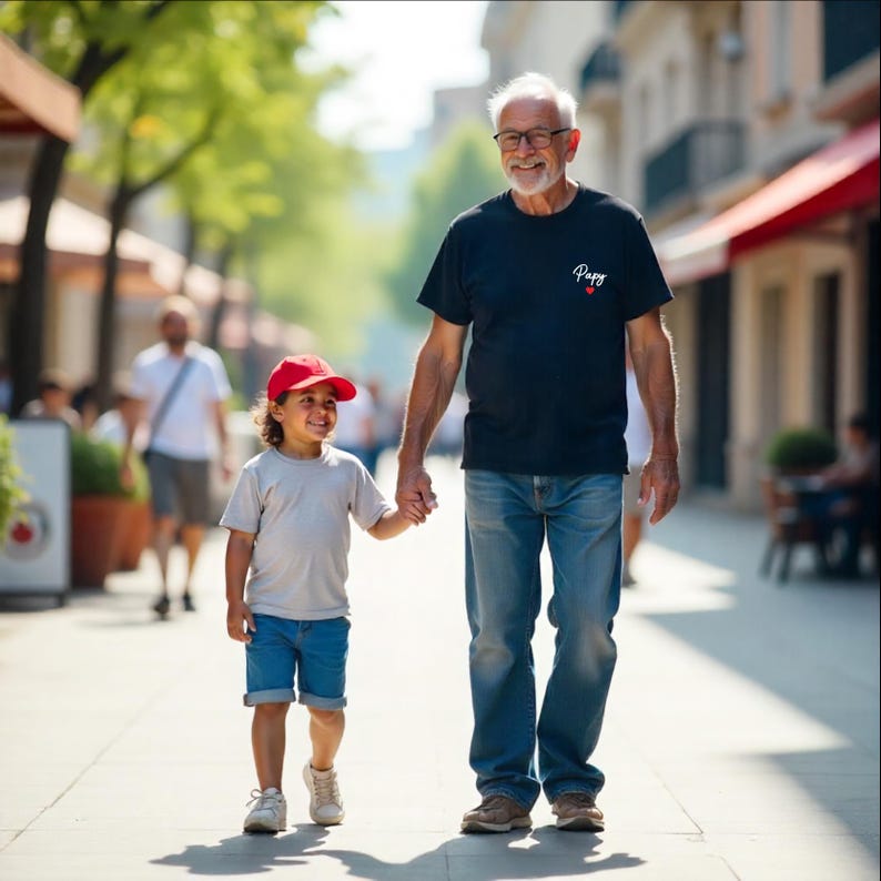 May include: A child and an older adult walk hand-in-hand down a sunny street. The child wears a red cap, gray shirt, and blue shorts. The adult wears a black shirt with "Papy" and a heart, blue jeans, and glasses.