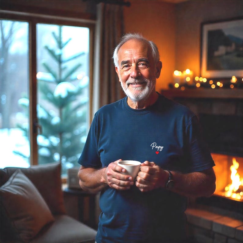 May include: A man wearing a navy blue t-shirt with the word "Papy" and a red heart. He is holding a white mug, smiling, and standing in front of a window and a fireplace.