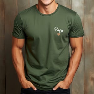 May include: Olive green t-shirt with the word "Papy" in cursive script and a small heart graphic. The shirt has short sleeves and a crew neck.