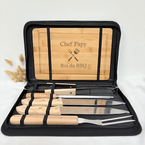 Op de afbeelding: Een zwarte etui met een bamboe snijplank en barbecuegereedschap. De plank is gegraveerd met "Chef Papy" en "Roi du BBQ". De set bevat een vleesvork, messen en tangen, allemaal met houten handvatten en roestvrijstalen bladen.