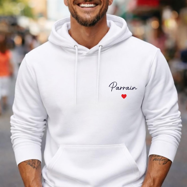 Puede incluir: Una sudadera con capucha blanca con un bolsillo delantero y un cord&oacute;n. La palabra "Parrain" est&aacute; impresa en el pecho en escritura negra, con un peque&ntilde;o coraz&oacute;n rojo debajo. La sudadera est&aacute; hecha de un material suave.