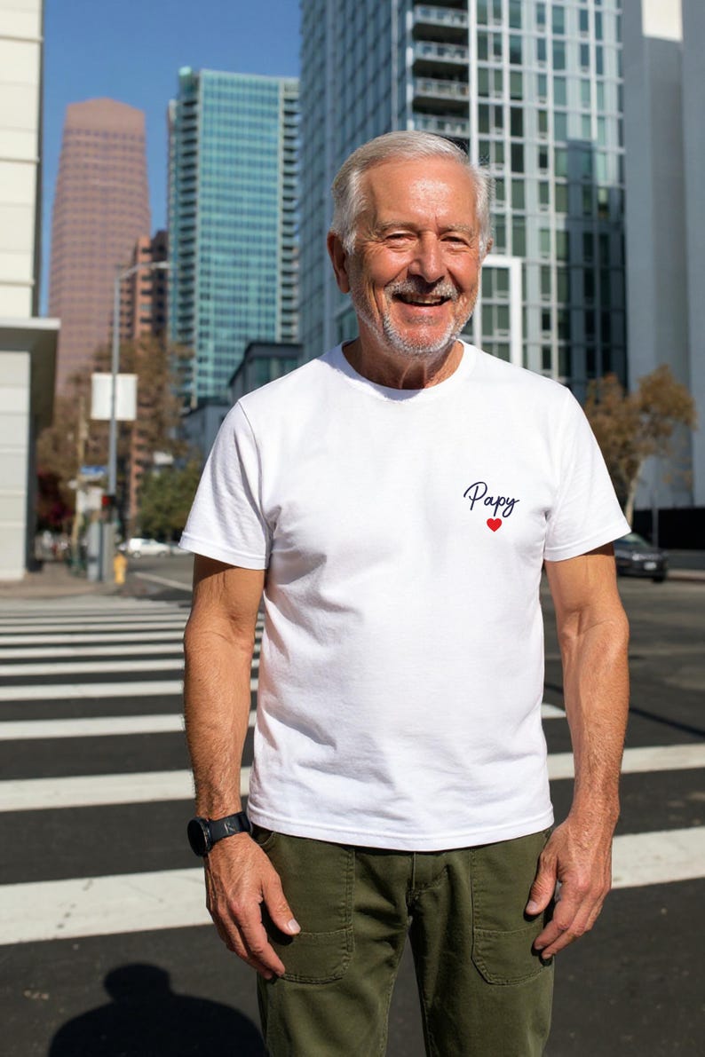 May include: White t-shirt with the word "Papy" in cursive and a small red heart. The shirt is worn with olive green pants and a black watch. The background features tall buildings and a crosswalk.