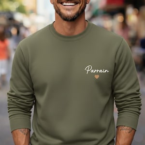 Puede incluir: Sudadera verde oliva con cuello redondo y la palabra "Parrain" en cursiva y un peque&ntilde;o coraz&oacute;n. La sudadera es de color liso y tiene mangas largas. El texto y el coraz&oacute;n son de color claro.