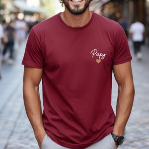 May include: A burgundy t-shirt with short sleeves. The shirt features the word "Papy" in cursive script, with a gold heart below it. The shirt is made of a soft material.