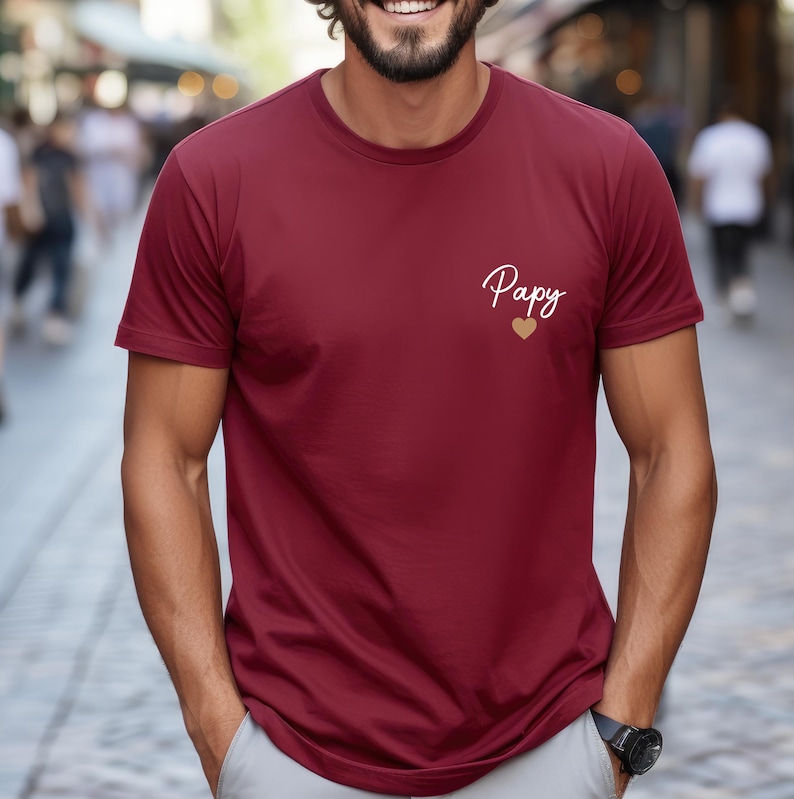 May include: A burgundy t-shirt with short sleeves. The shirt features the word "Papy" in cursive script, with a gold heart below it. The shirt is made of a soft material.