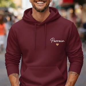 Puede incluir: Una sudadera con capucha burdeos con un bolsillo delantero y una capucha con cord&oacute;n. La palabra "Parrain" est&aacute; escrita en cursiva en el pecho, acompa&ntilde;ada de un peque&ntilde;o coraz&oacute;n dorado.
