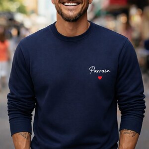 Puede incluir: Sudadera azul marino con la palabra "Parrain" en escritura blanca y un peque&ntilde;o coraz&oacute;n rojo. La sudadera de cuello redondo es de color liso y tiene mangas largas.