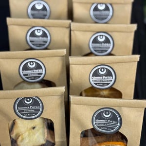 May include: A collection of brown paper bags with clear windows, each containing dried botanicals. The bags are labeled with black and white circular stickers that read "Simmer Pot Kit" and include the words "Cleansing" or "Blessings & Protection".