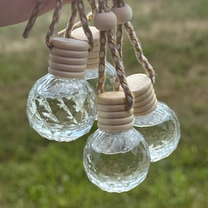 May include: A group of small, clear glass bottles with a faceted, diamond-like texture. Each bottle features a light-colored wooden cap and is suspended by a braided beige cord interwoven with thin gold metallic threads and a wooden bead.