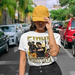 May include: A white t-shirt with a graphic print of a group of people. The print features the text "Sidhu Moose Wala" in gold lettering. The person wearing the shirt is also wearing a yellow baseball cap.