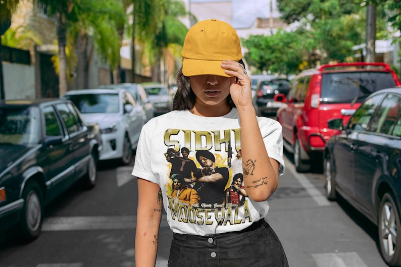May include: A white t-shirt with a graphic print of a group of people. The print features the text "Sidhu Moose Wala" in gold lettering. The person wearing the shirt is also wearing a yellow baseball cap.