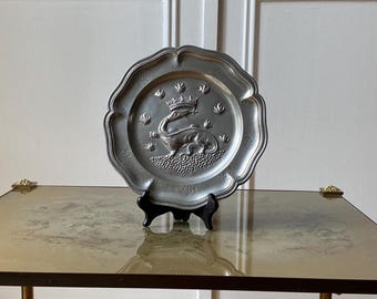May include: A silver decorative plate with a raised design of a crowned dragon. The plate is on a black stand and a glass table.