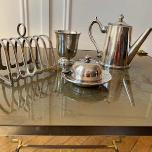 Peut inclure: Un ensemble de service en argent sur une table en verre. L'ensemble comprend un beurrier, une théière, un porte-toast et une tasse en argent.