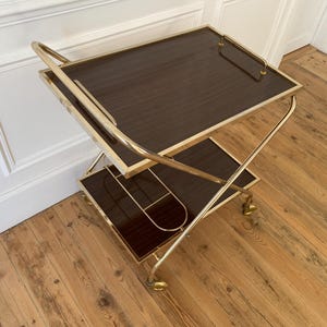 Puede incluir: Un carrito de bar vintage con estructura de metal dorado y dos estantes rectangulares de color marrón oscuro. El carrito tiene asas curvas y ruedas pequeñas. Los estantes tienen un aspecto de veta de madera oscura.
