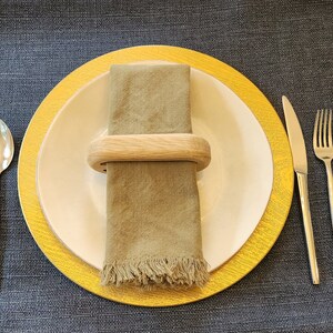 Könnte beinhalten: Ein weißer Speiseteller mit goldfarbenem Rand liegt auf einem goldfarbenen Unterteller. Eine grüne Leinen-Serviette wird von einem Serviettenring aus Holz gehalten. Zwei silberne Löffel und drei silberne Gabeln liegen auf einer dunkelblauen Tischdecke.
