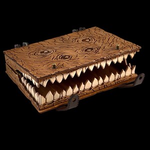 May include: A wooden box with a carved monster face on the lid. The box is open, revealing a set of sharp teeth inside. The box is made of light brown wood and has a dark brown stain.