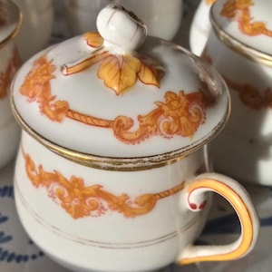May include: Several white porcelain sugar bowls with lids, featuring orange floral designs and gold edging. Each bowl has a small handle and rests on a decorative blue and white patterned plate. The lids are topped with a small white knob.